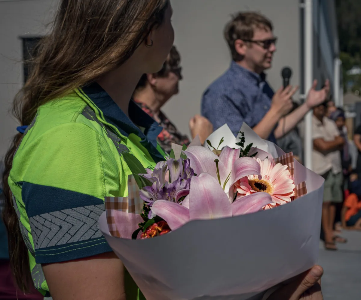 Close-up of a bouquet held by a woman in a high-vis shirt while a speaker addresses guests at a Habitat Nelson dedication event