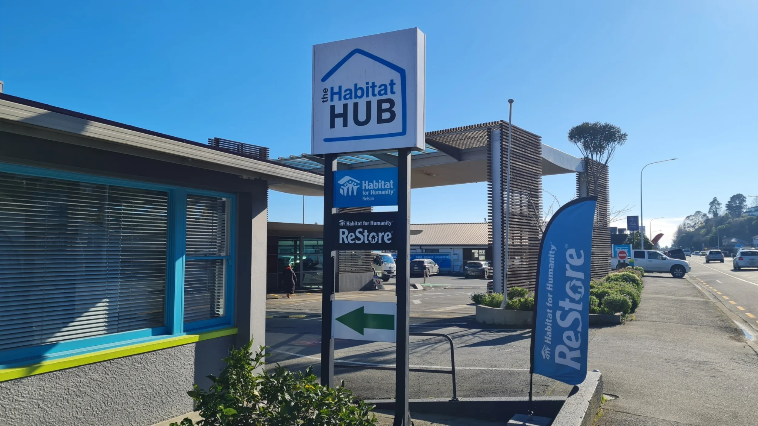 Entrance to the Habitat Hub in Nelson, featuring prominent Habitat Hub and Habitat for Humanity ReStore signage, a blue ReStore feather flag, and the driveway leading into the site on a clear sunny day.